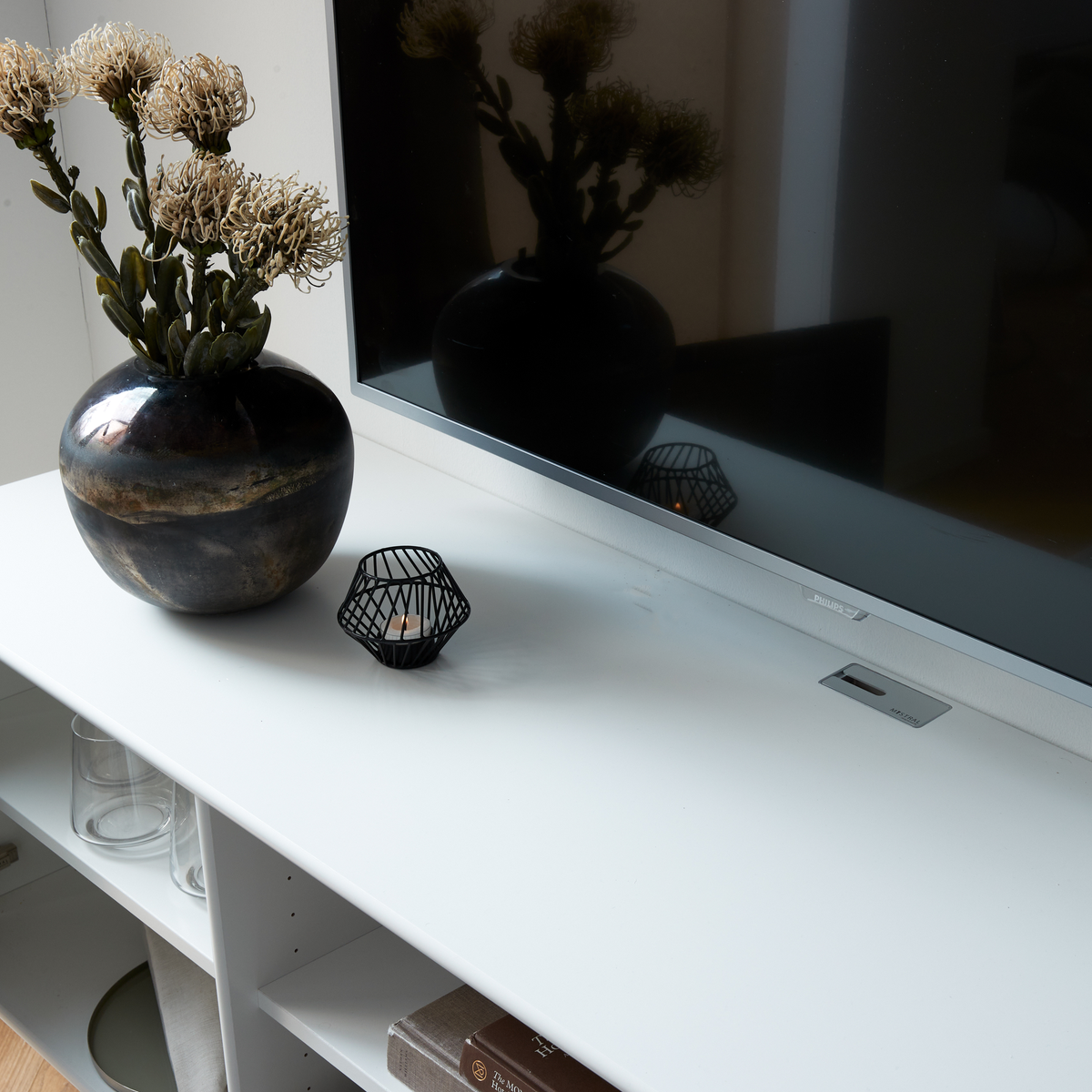 Hvid tv-bænk med vase med tørrede blomster, tændt fyrfadsstage og et Philips‑tv.