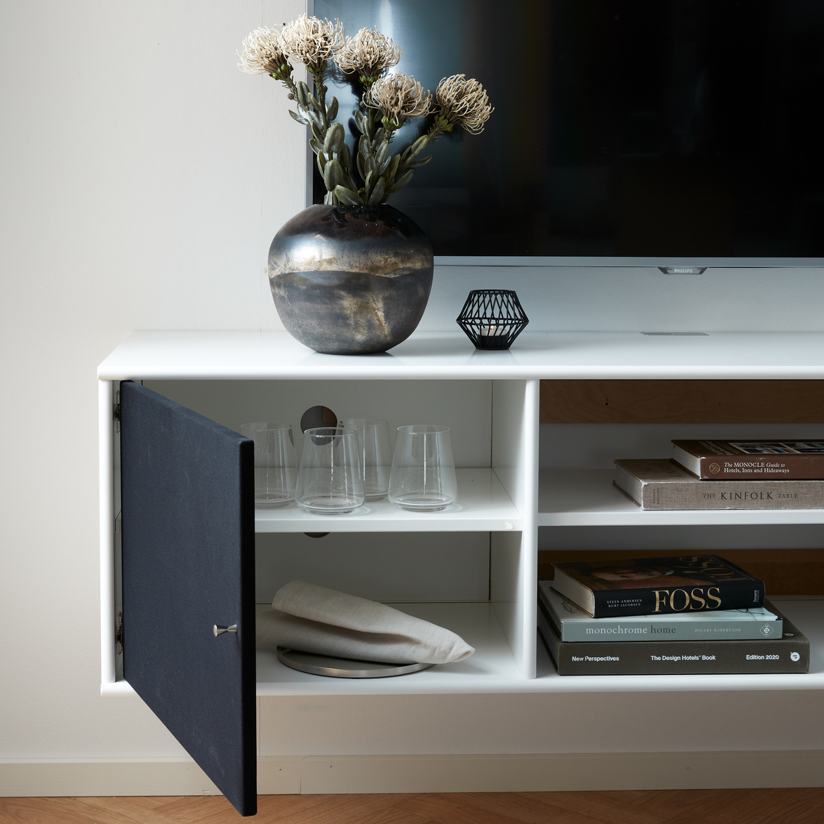 Hvid mediabænk med TV, vase med tørrede blomster og åben låge med glas; ved siden af ligger en stak designbøger: The Kinfolk Table, Monochrome Home, The Design Hotels Book, New Perspectives (Edition 2020) og Foss.