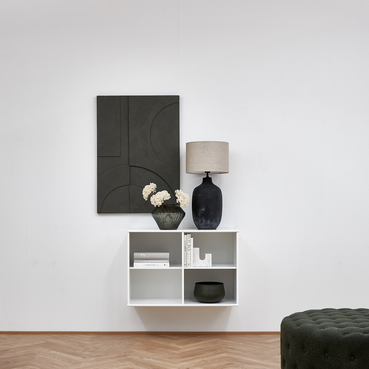 Minimalistisk stue med vægmonteret hvid reol, bordlampe med beige skærm, mørk vase med hvide blomster og sort geometrisk vægkunst.