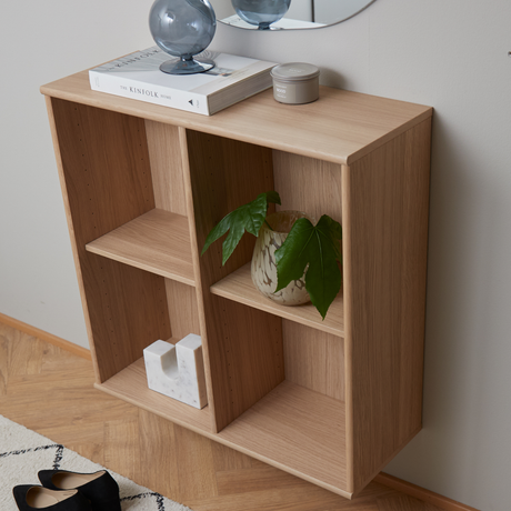 Vægmonteret reol i lyst egetræ med bogen The Kinfolk Home, en grå glasdekoration og en lille lysdåse ovenpå samt en grøn plante i krukke og et hvidt marmordekor i rummene.