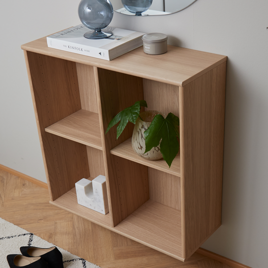 Vægmonteret reol i lyst egetræ med bogen The Kinfolk Home, en grå glasdekoration og en lille lysdåse ovenpå samt en grøn plante i krukke og et hvidt marmordekor i rummene.