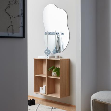 Minimalistisk entré med vægmonteret egereol under et organisk formet spejl, stylet med blå lysestager, en bog og en lille plante.