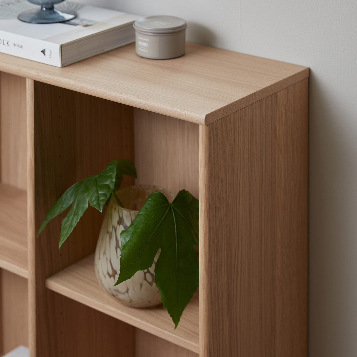 Lys træreol med grøn plante i mønstret vase; ovenpå ligger en bog med titlen HOME og en grå dåse mærket MOOD.