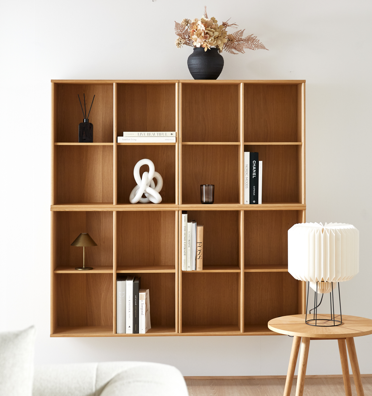 Væghængt reol i træ med bøger, CHANEL‑bog, skulpturel hvid knude, sort vase med tørrede blomster samt en lille bordlampe og en plisseret bordlampe på et sidebord.