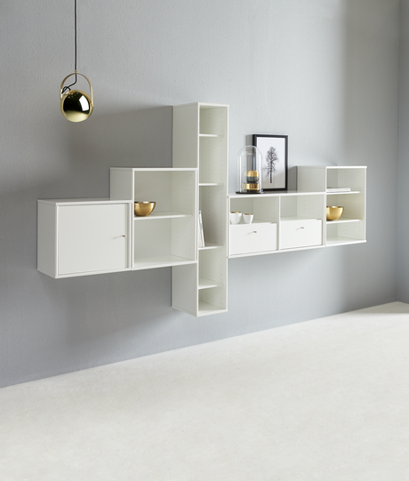 Vægmonterede hvide reoler mod en grå væg med en messingfarvet pendellampe, guldfarvede skåle, bøger, glaskuppel og indramet billede; luftig, minimalistisk styling.