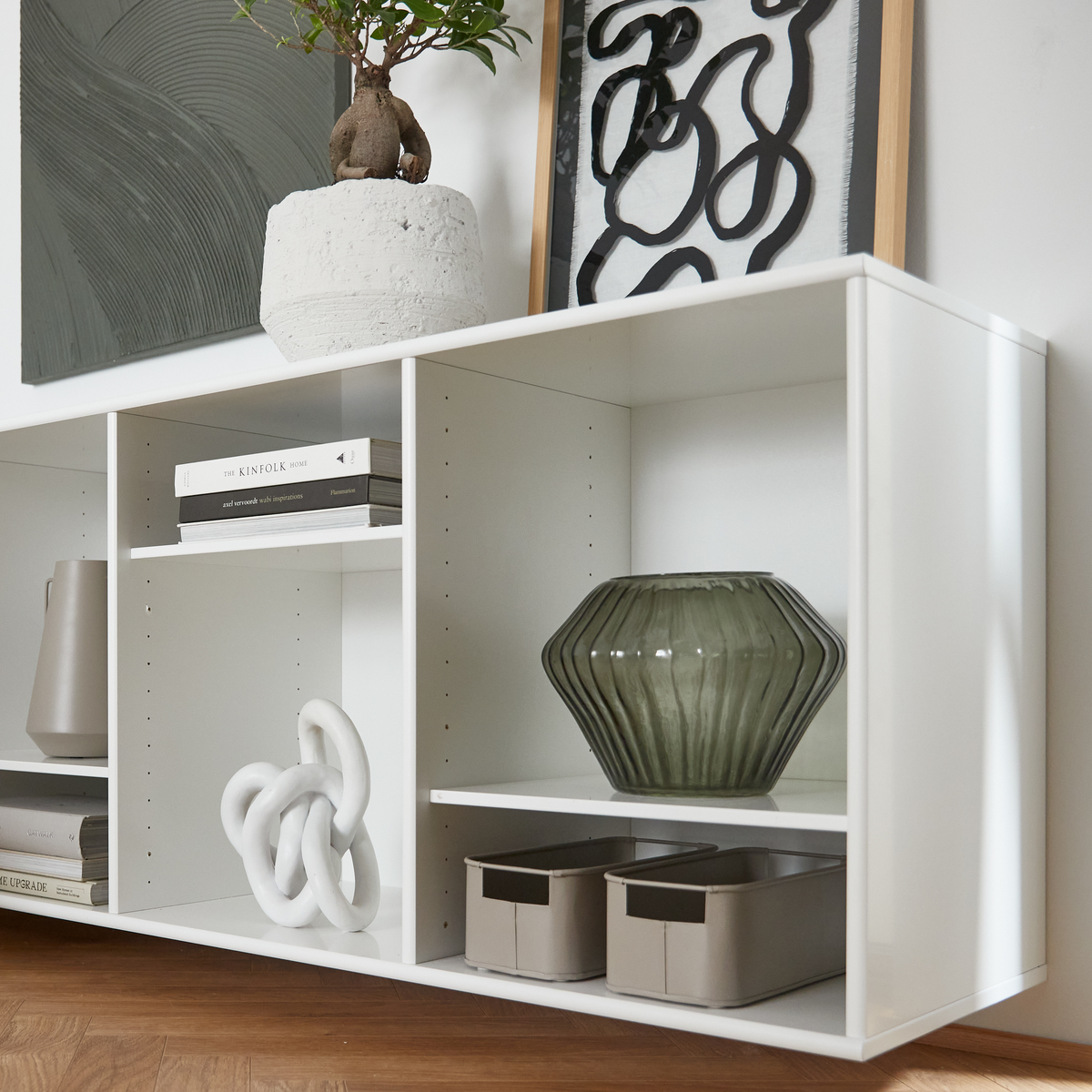 Hvid reol med grøn riflet glasvase, abstrakt kunst og potteplante ovenpå, bøger inklusiv The Kinfolk Home, skulpturel knude og beige opbevaringskasser.