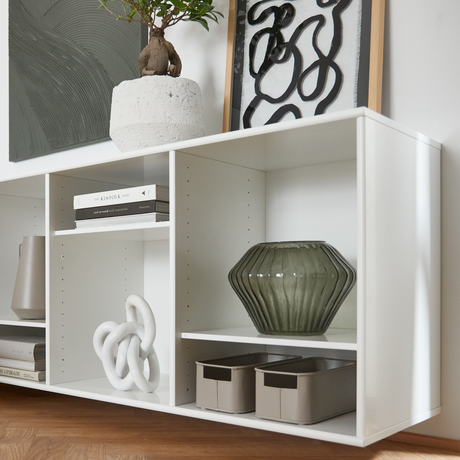 Hvid reol med grøn riflet glasvase, abstrakt kunst og potteplante ovenpå, bøger inklusiv The Kinfolk Home, skulpturel knude og beige opbevaringskasser.