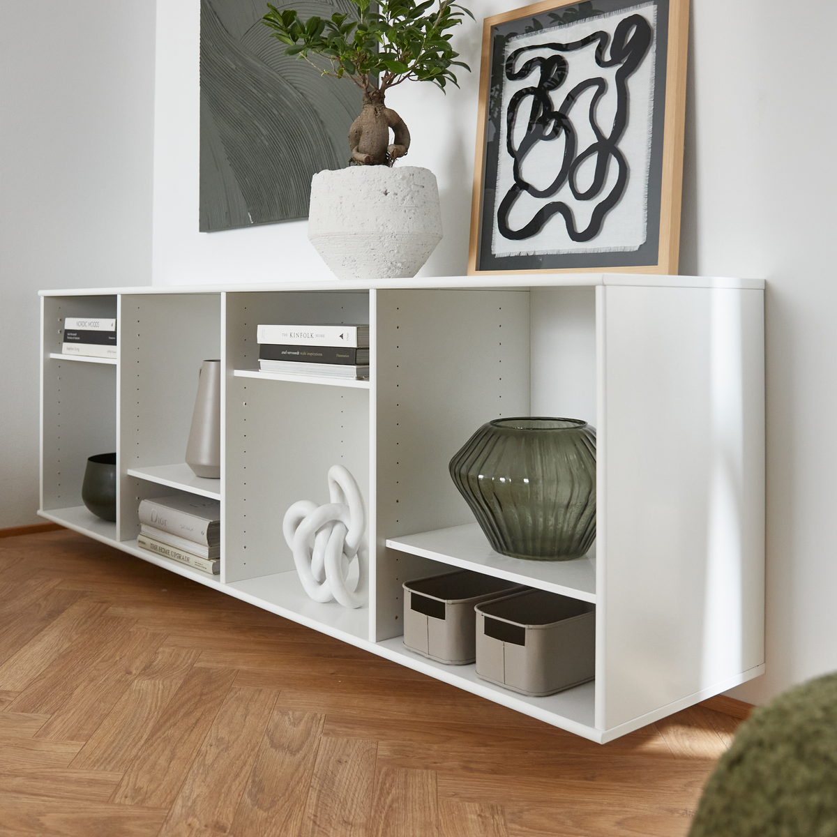 Hvid lav reol med åbne rum, stylet med bøger, grøn glasvase, to opbevaringskasser og bonsai i potte; abstrakt kunst op ad væggen.