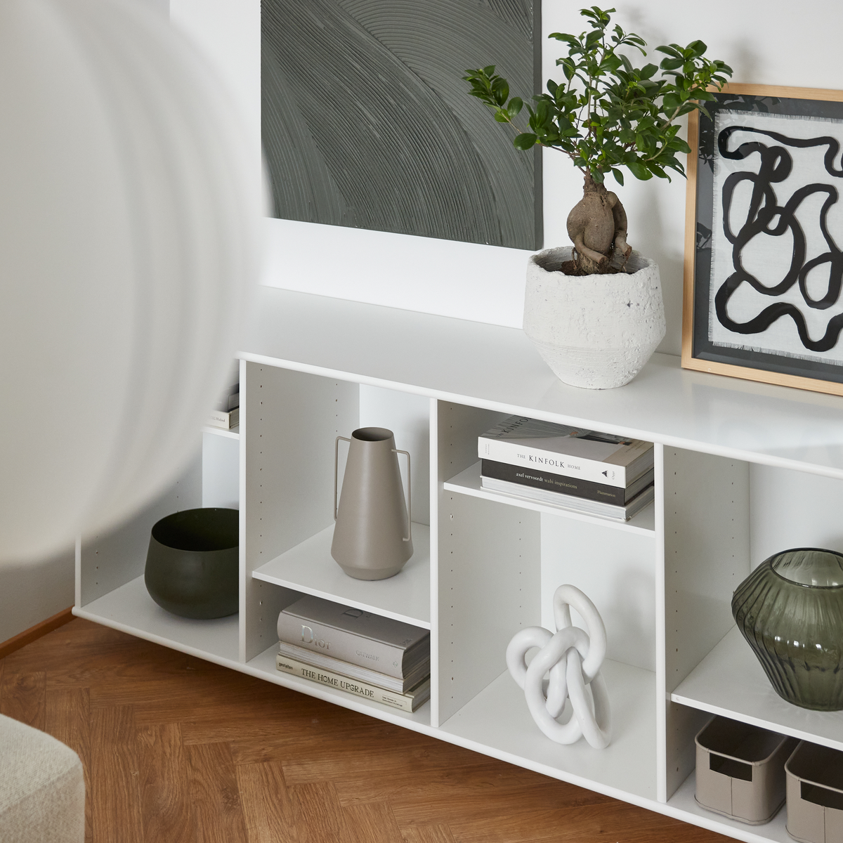 Hvid reol med kunst, grøn plante i lys krukke, dekorative vaser og skulptur; bøgerne The Kinfolk Home og The Home Upgrade ligger på hylderne.