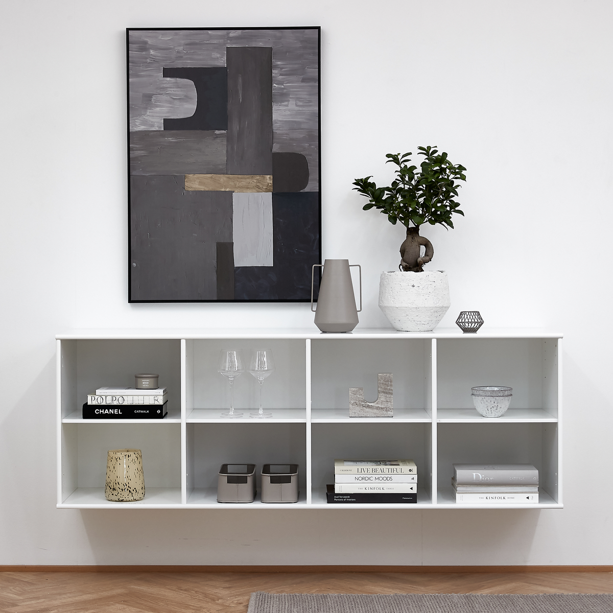 Hvid væghængt reol med dekoration; plante i potte, grå kande, glas, lysstage og abstrakt kunst på væggen. Bøger inkl. Chanel, Nordic Moods, Live Beautifully og The Kinfolk.
