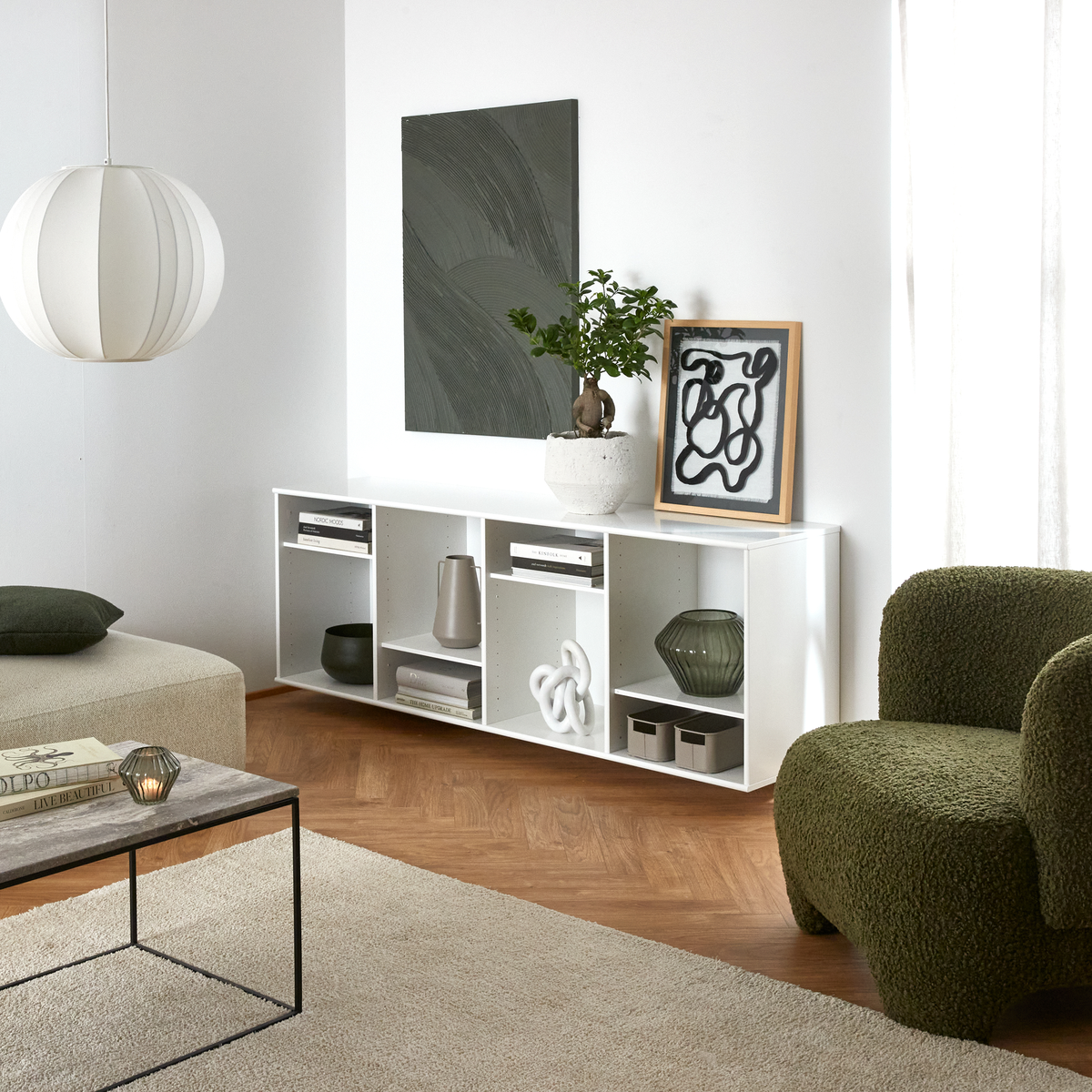 Lyst stue med hvid lav reol, grøn lænestol, pendel, abstrakt kunst, plante i krukke og bogen Dior på sofabordet.