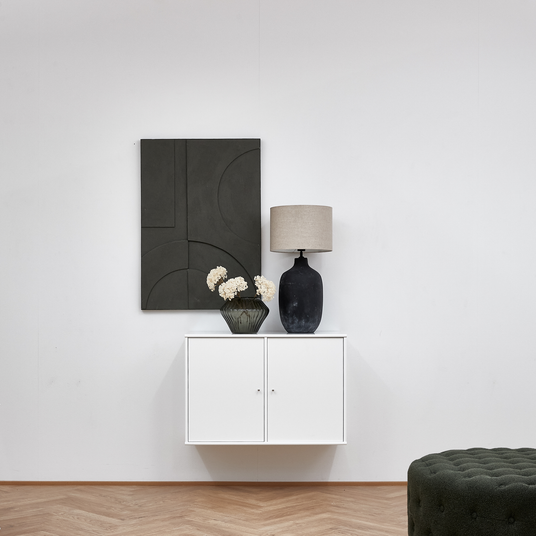 Vægmonteret hvidt skab med sort bordlampe, mørkegrøn vase med lyse blomster og mørk geometrisk vægkunst over; grøn puf i forgrunden.