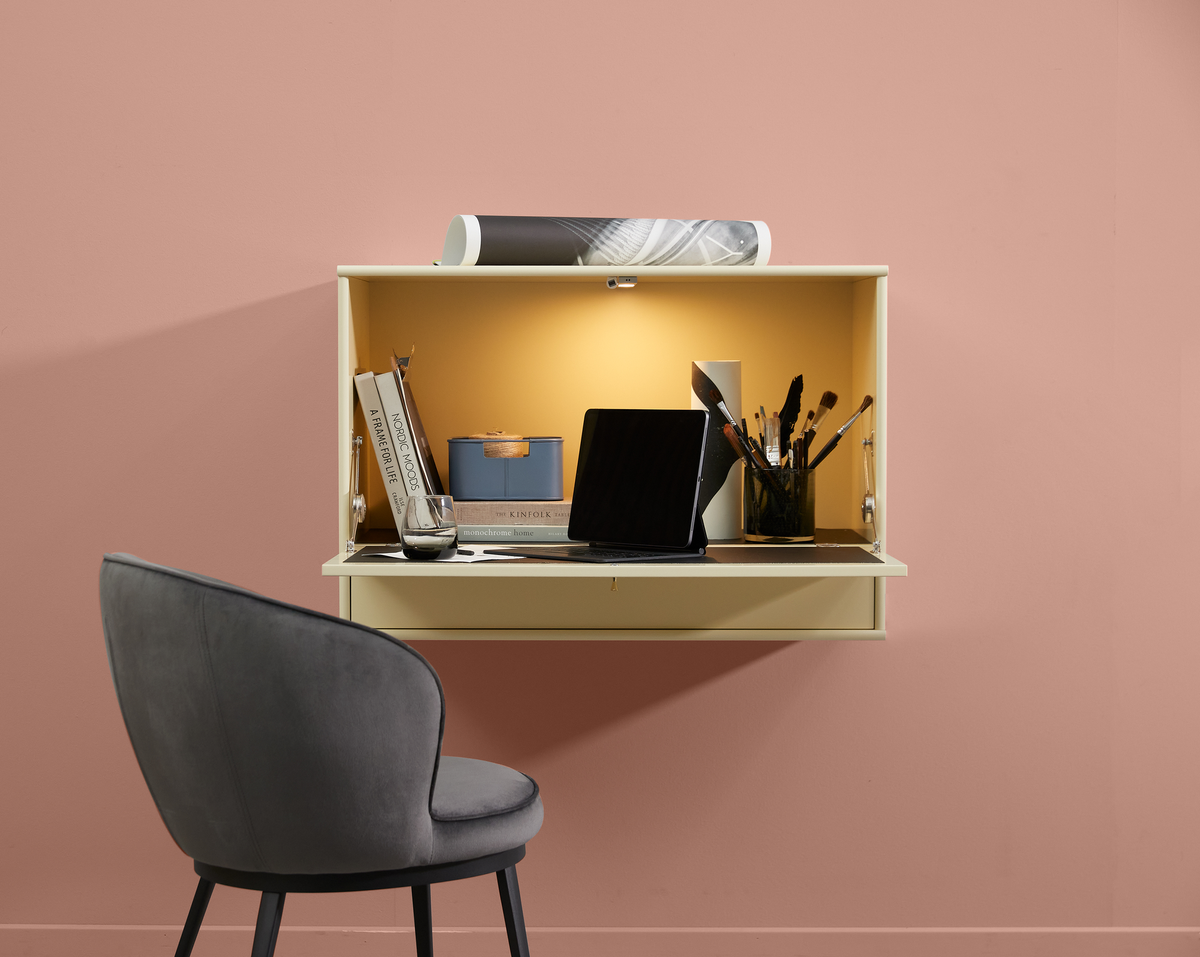 Væghængt lille arbejdsplads med tablet, bøgerne A Frame for Life, Nordic Moods og The Kinfolk Home, pensler i glas og blå boks, varm belysning og grå stol foran rosa væg.