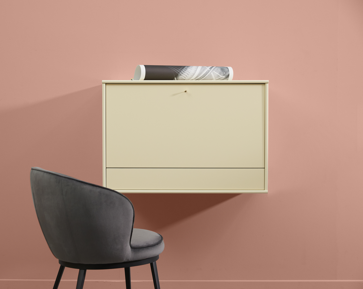 Beige væghængt skab på rosa væg med grå polstret stol ved siden af og en sammenrullet plakat ovenpå.