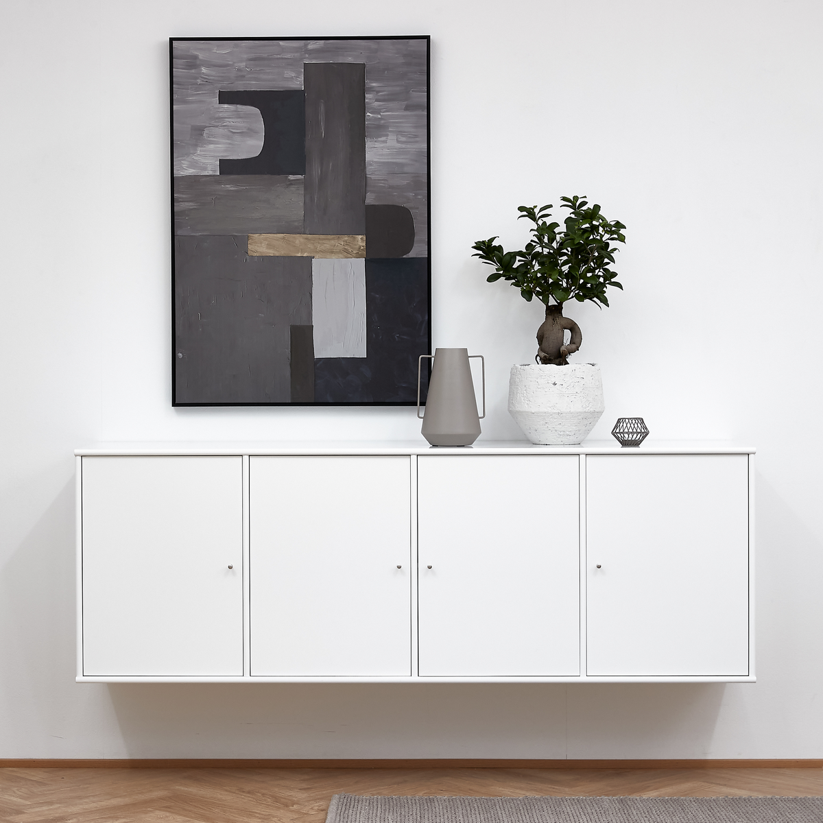 Vægmonteret hvid skænk med abstrakt gråt kunstværk ovenover, grå vase, lille dekoration og grøn plante i hvid krukke i en lys, minimalistisk indretning.