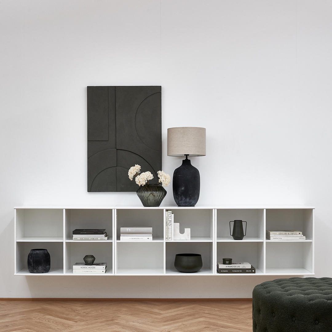 Minimalistisk stue med vægmonteret hvid reol, sort bordlampe, vase med lyse blomster, bøger og mørkegrå vægkunst.