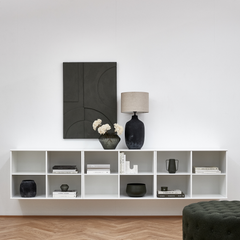 Minimalistisk stue med vægmonteret hvid reol, sort bordlampe, vase med lyse blomster, bøger og mørkegrå vægkunst.