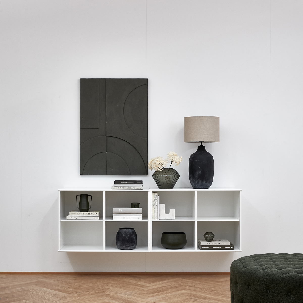 Minimalistisk reol med sorte og grønne vaser, beige bordlampe, grafisk mørkt kunstværk og bøger, bl.a. ”A Frame for Life” og ”Lindform”, i en lys stue.