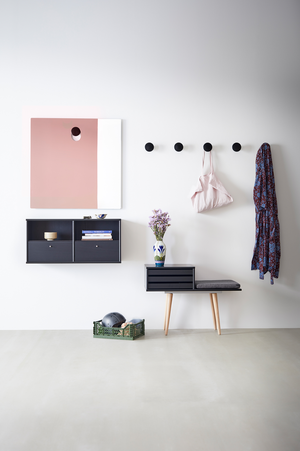 Lys entré med sorte runde vægknager, lille sort bænk med grå pude, væghylder, rosa‑hvidt spejl og en vase med lilla blomster samt en grøn kasse på gulvet.