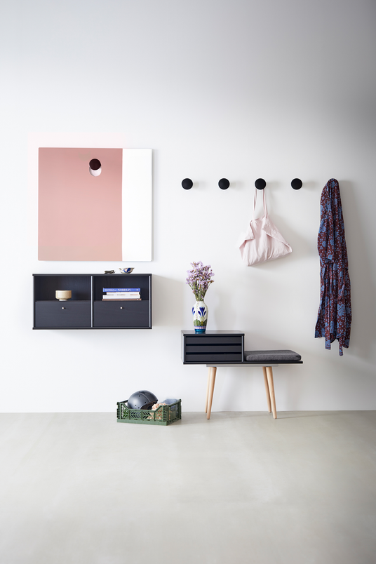 Lys entré med sorte runde vægknager, lille sort bænk med grå pude, væghylder, rosa‑hvidt spejl og en vase med lilla blomster samt en grøn kasse på gulvet.