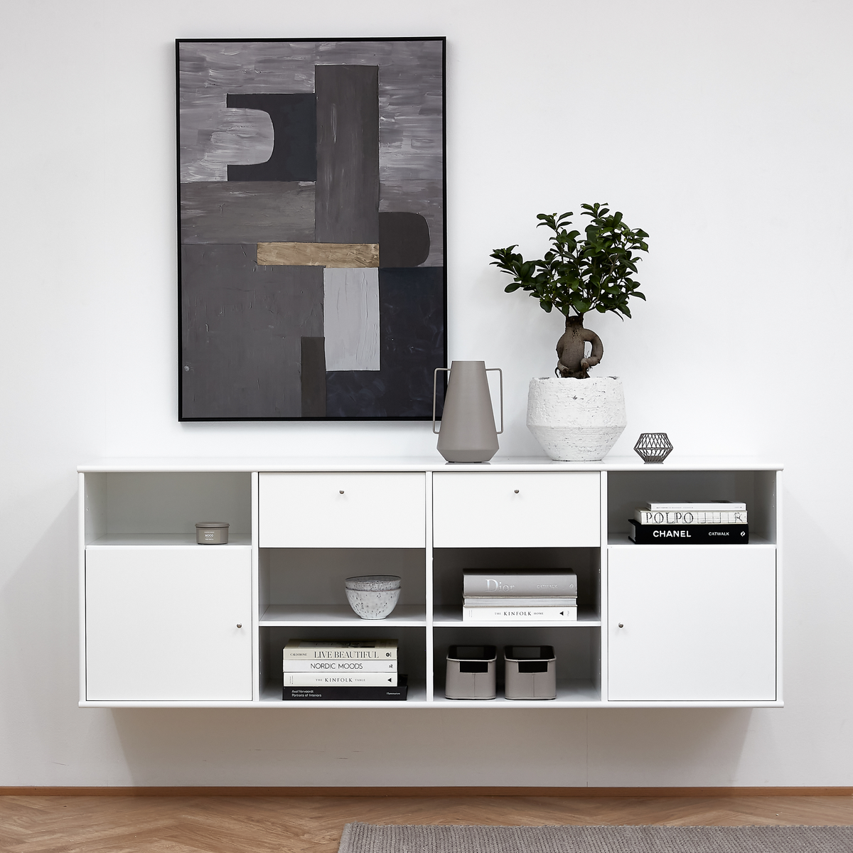 Minimalistisk hvid væghængt skænk med åbne hylder med bøger – Chanel, Kinfolk, Live Beautiful, Polpo, Dior – samt skål og opbevaringsbokse; over hænger et abstrakt gråt billede, og på toppen står en grøn plante i hvid potte og en grå kande.