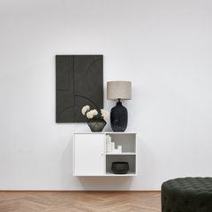 Minimalistisk stue med vægmonteret hvidt skab, sort keramisk bordlampe med beige skærm, mørk glasvase med hvide blomster og sort geometrisk vægkunst; sildebensparket og grøn skammel.