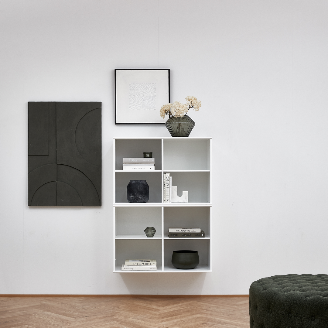 Væghængt hvid reol med bøger og dekoration; grøn glasvase med tørrede blomster ovenpå, to vægbilleder og en mørkegrøn ottoman; bogen “A Frame of Life” findes i reolen.