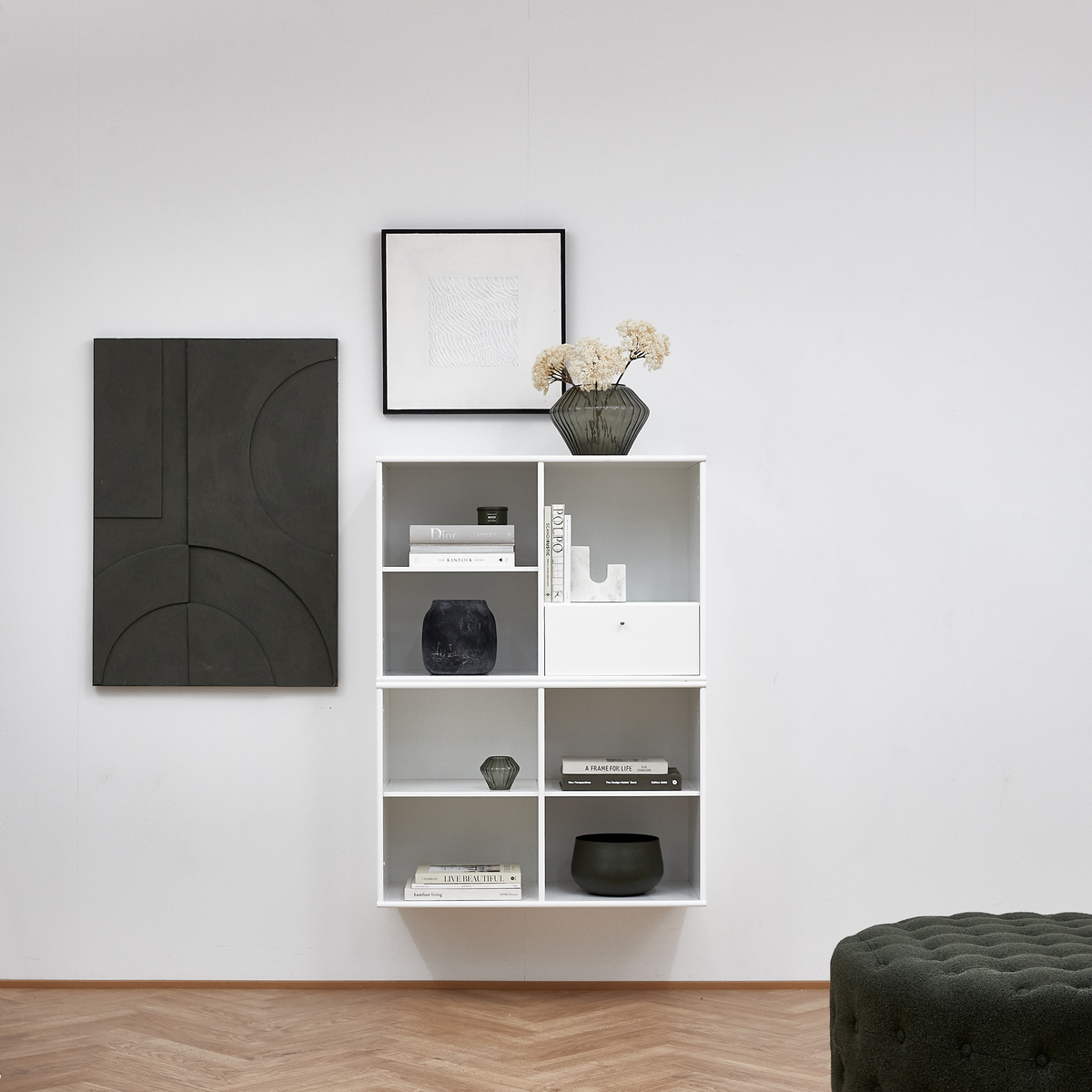 Hvid væghængt reol med vaser og bøger, bl.a. "A FRAME FOR LIFE", på lys væg; to billeder over og grøn ottoman i forgrunden.