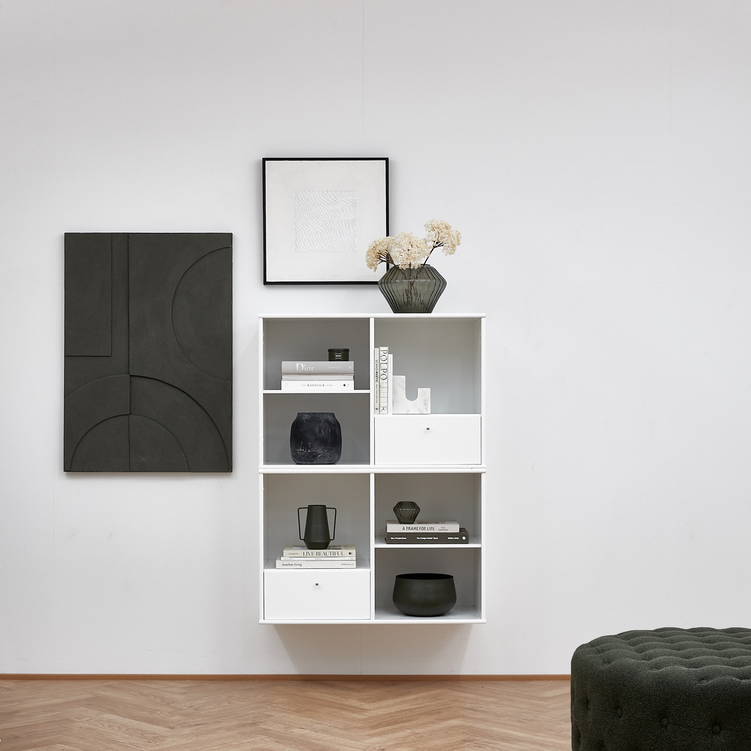 Vægmonteret hvid reol med bøger og keramik; titlerne ”Dior” og ”A FRAME APOLLO” er synlige; grøn glasvase med tørrede blomster ovenpå; to grafiske vægbilleder og en mørkegrøn puf i forgrunden.