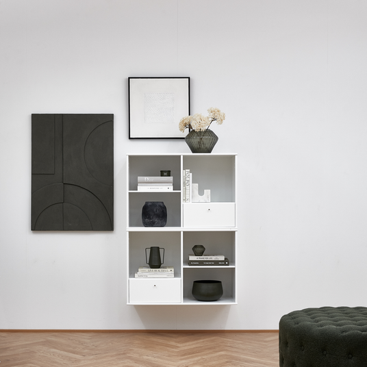 Vægmonteret hvid reol med bøger og keramik; titlerne ”Dior” og ”A FRAME APOLLO” er synlige; grøn glasvase med tørrede blomster ovenpå; to grafiske vægbilleder og en mørkegrøn puf i forgrunden.