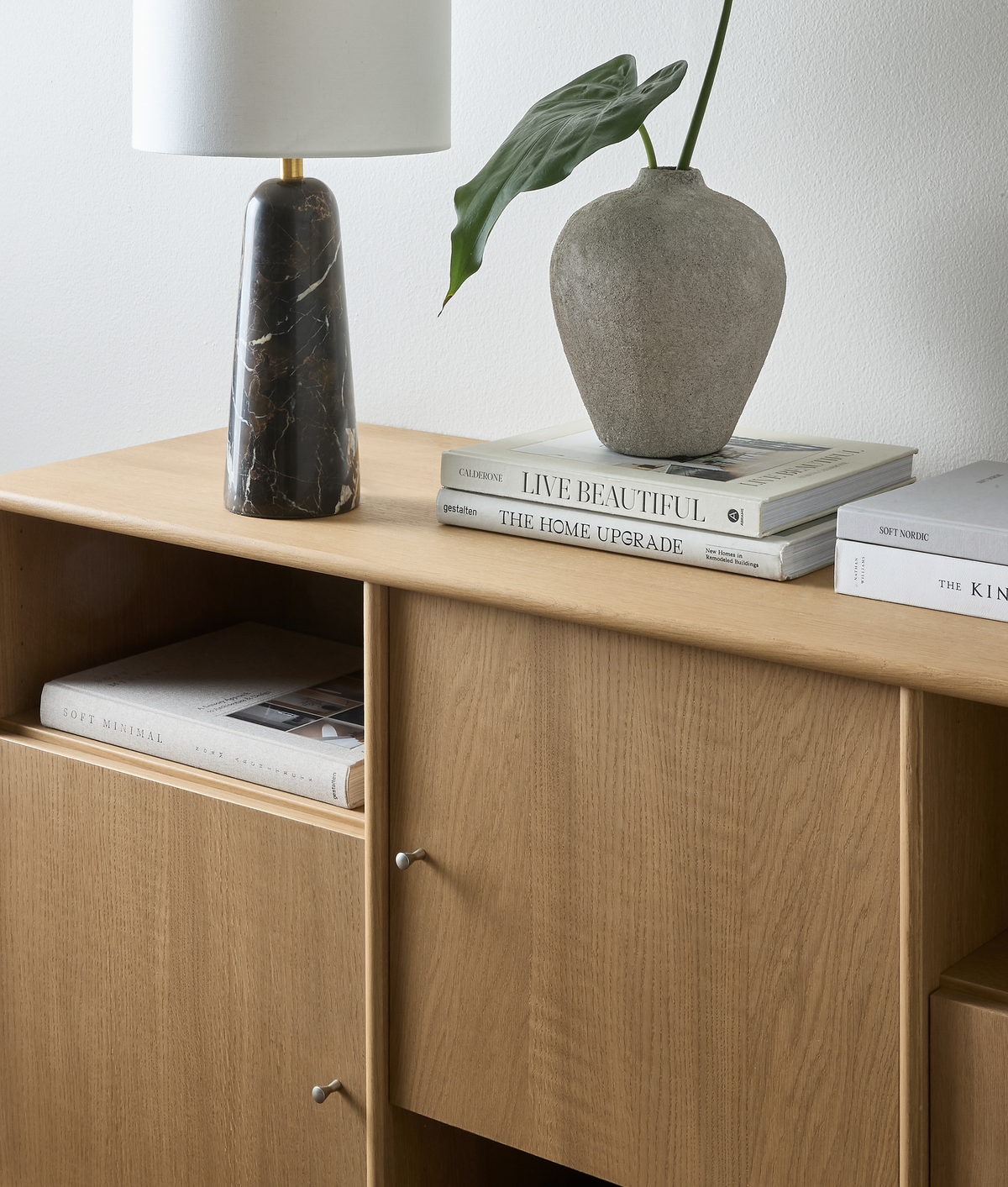 Træskænk med marmorbordlampe, grå vase med grøn bladstilk og stakkede bøger med titlerne ”LIVE BEAUTIFUL”, ”THE HOME UPGRADE”, ”SOFT MINIMAL” samt ”THE KIN…”.