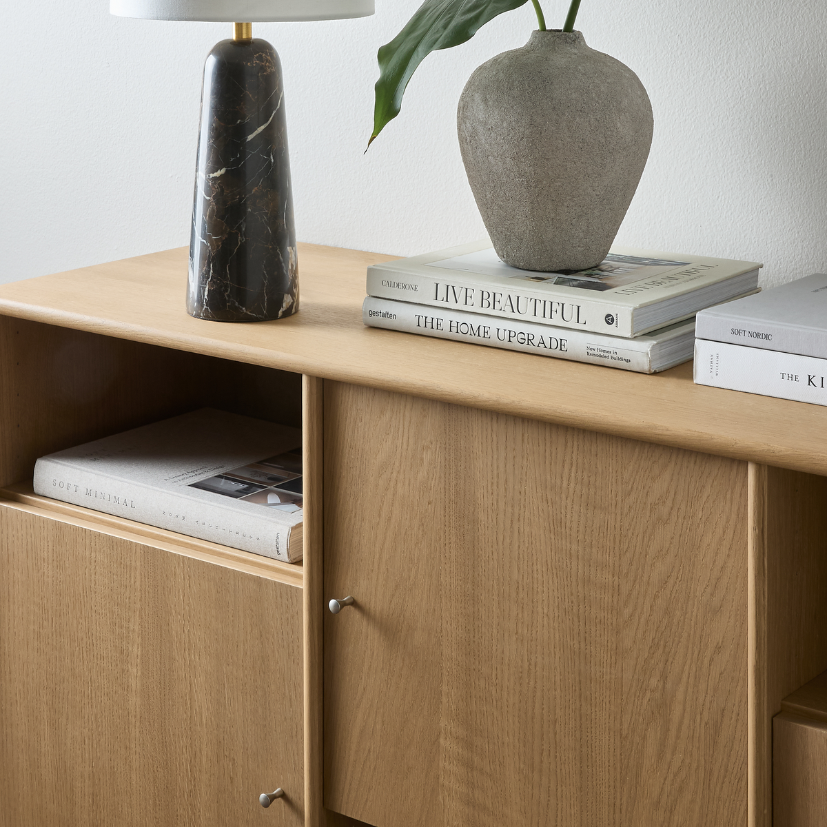 Nærbillede af et egeskab med sort marmorbordlampe, grå keramisk vase med grønt blad og stablede bøger: Live Beautiful og The Home Upgrade.