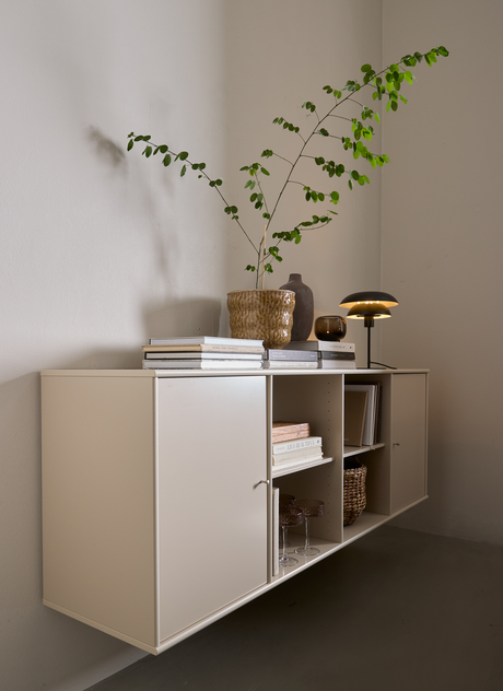 Beige væghængt skab med åbne rum, bøger, en keramikvase med grønne grene og en tænd bordlampe.