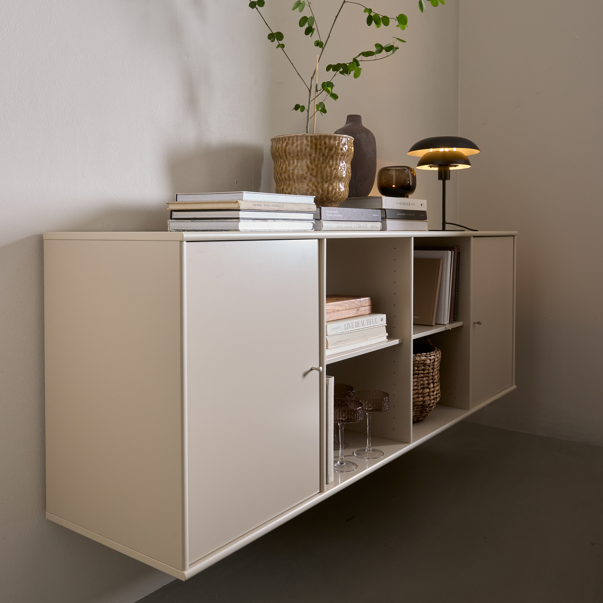 Beige vægmonteret skab med åbne hylder, bøger, bordlampe og krukke med grøn gren.