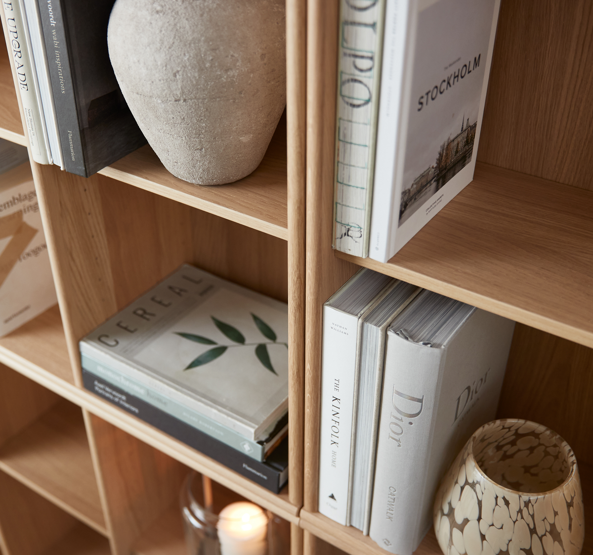 Reol i træ med designbøger som Stockholm, The Kinfolk Home, Dior og Cereal, sammen med en stenvase, et mønstret glas og et tændt lys.