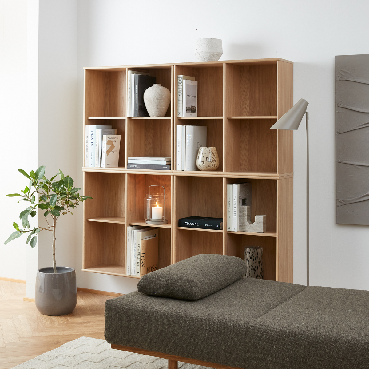 Lyst stue med reol i eg med bøger som PRADA og CHANEL, vaser og en tænd lyslygte, ved siden af en grå daybed, gulvlampe og potteplante.