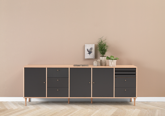 Skænk i lyst træ med mørkegrå fronter, stylet med små potteplanter og dekorationer mod beige væg.