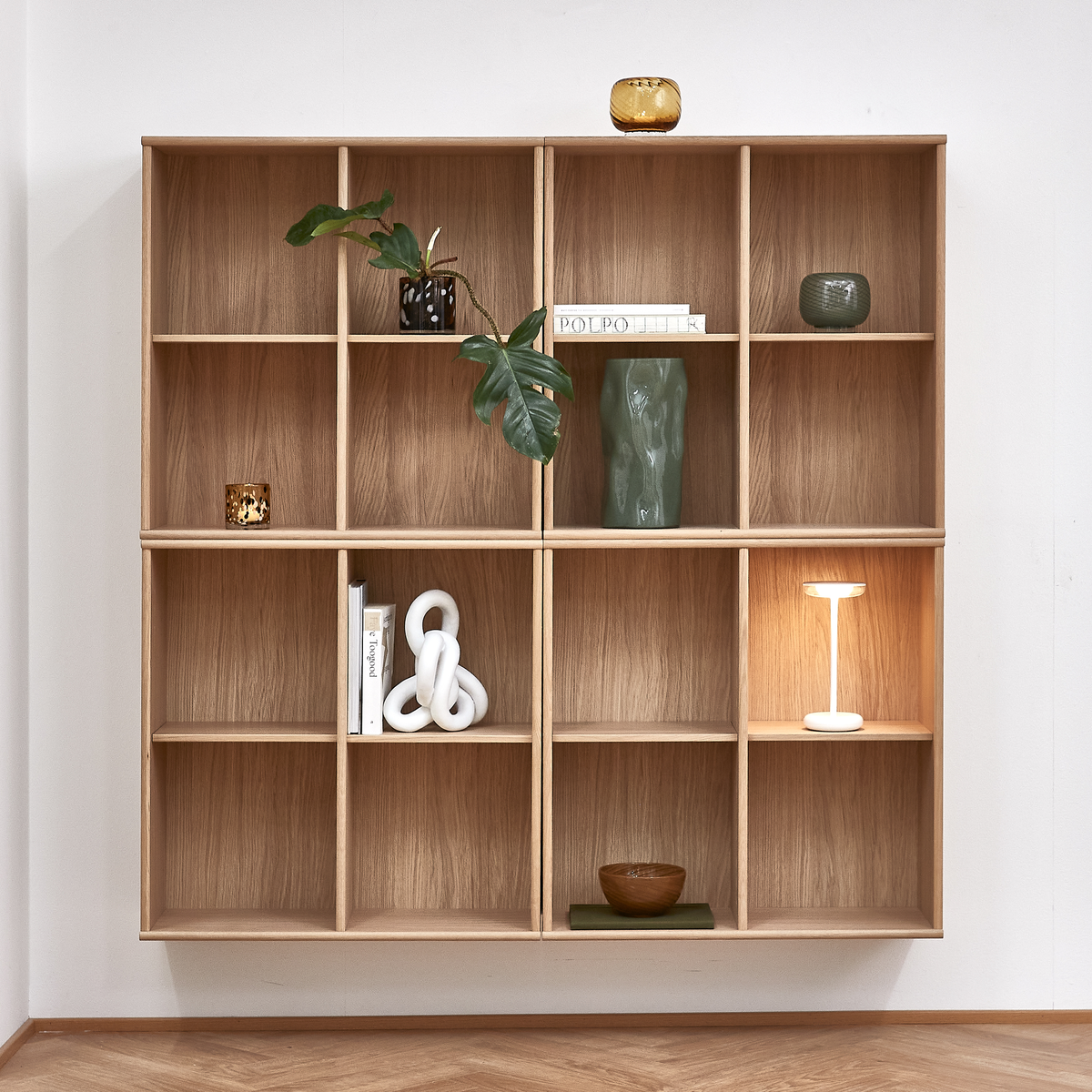 Vægmonteret reol i træ med bøger, plante, grøn vase, små skåle og en hvid trådløs bordlampe; en bog med titlen POLPO ses.