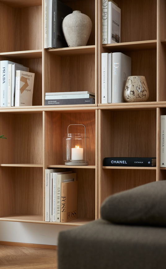 Nærbillede af en åben reol i lyst træ med interiørbøger og dekorationer; titler som Prada, Dior, Chanel Catwalk, The Kinfolk Home og FOSS ses, samt en tænd glaslanterne med stearinlys.