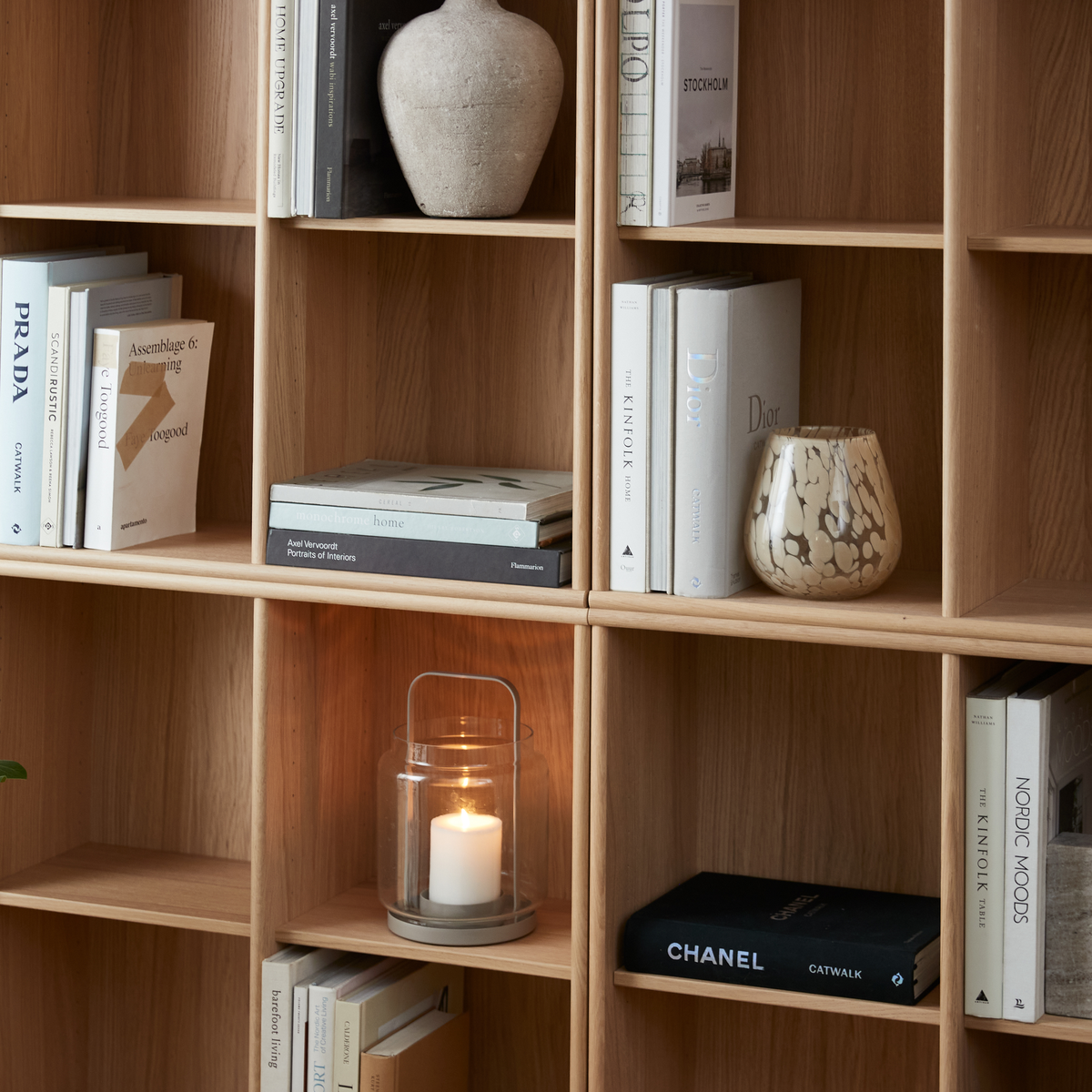Åben reol i lys eg med designbøger som Prada, Dior, Chanel, Kinfolk og Nordic Moods, en tændt lyslygte og en keramikvase.