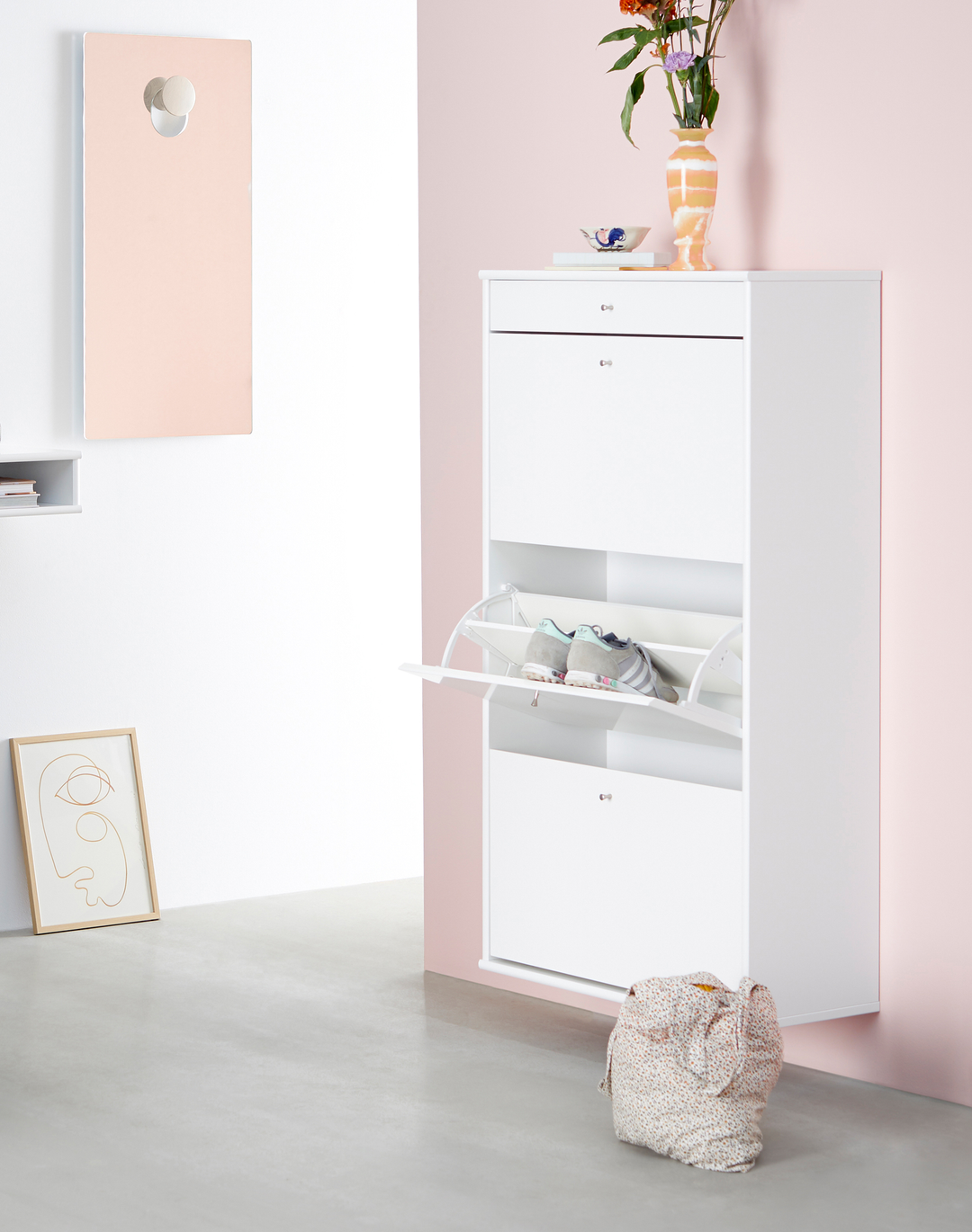 Vægmonteret hvidt skoskab med klaplåger, en låge åben med sko, mod rosa væg og en vase med blomster ovenpå.