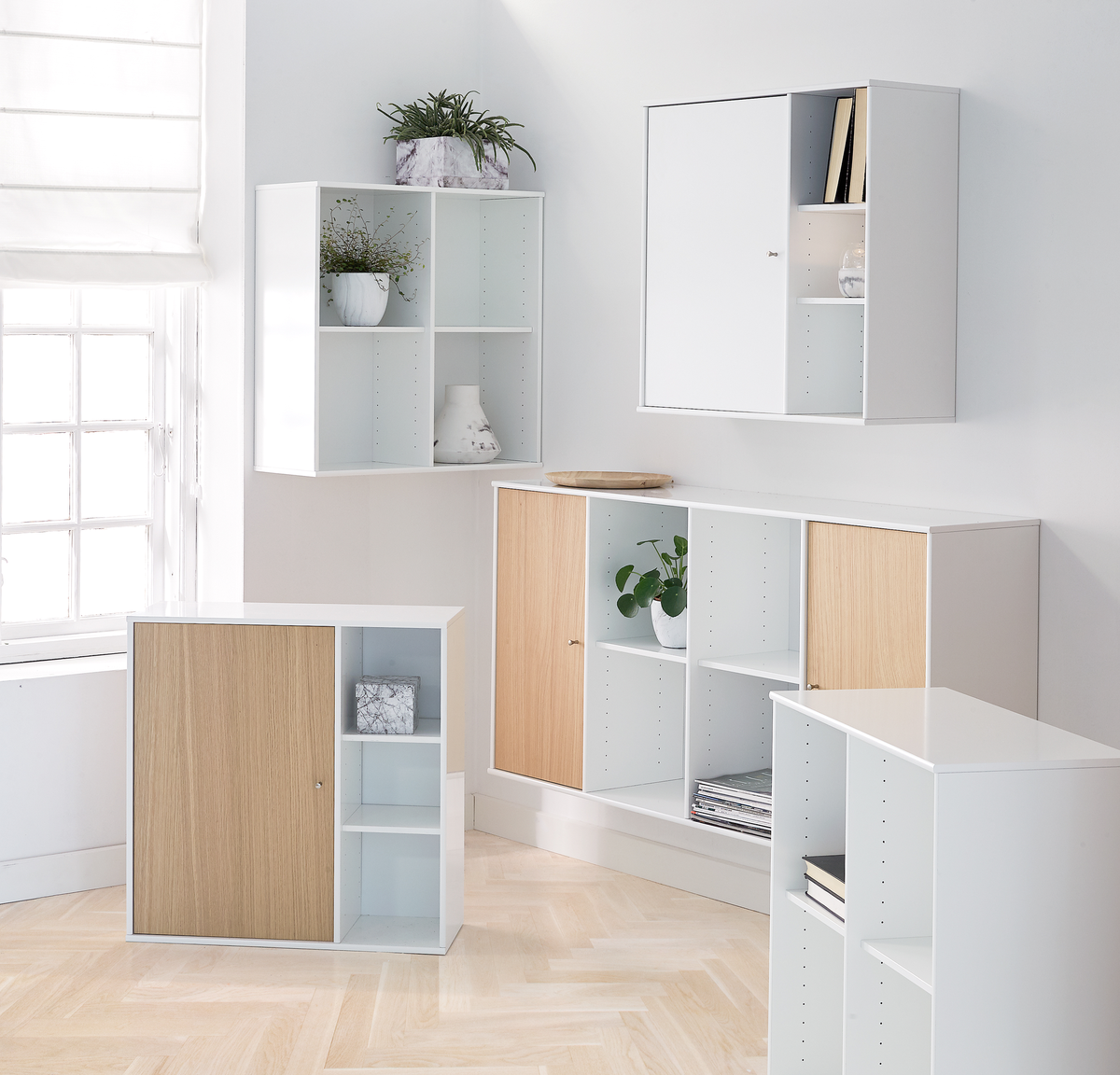 Lyse hvide opbevaringsmoduler med skydelåger i egetone, stylet med planter, bøger og keramik i et sollyst rum.