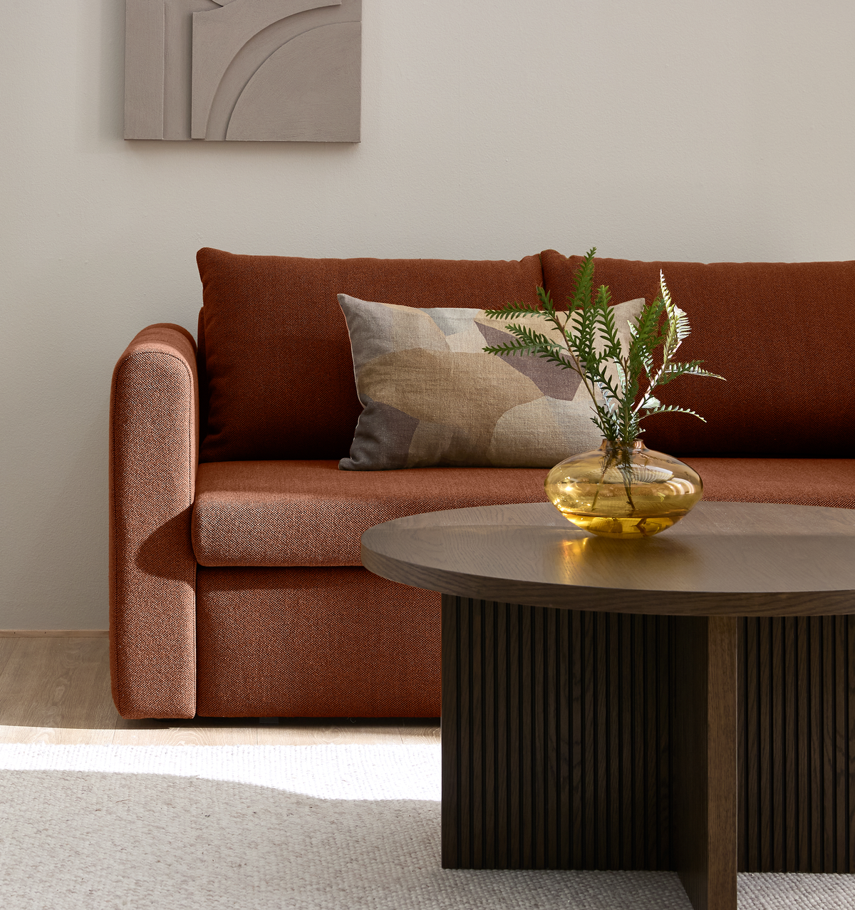 Rustorange sofa med mønstret pude, mørkt rundt sofabord og gul glasvase med grønne kviste i en lys stue.