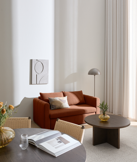 Rolig skandinavisk stue og spiseplads med rustfarvet sofa, rundt mørkt sofabord med glasvase, gulvlampe og lyse gardiner; spisebord i forgrunden med åben bog i blødt dagslys.