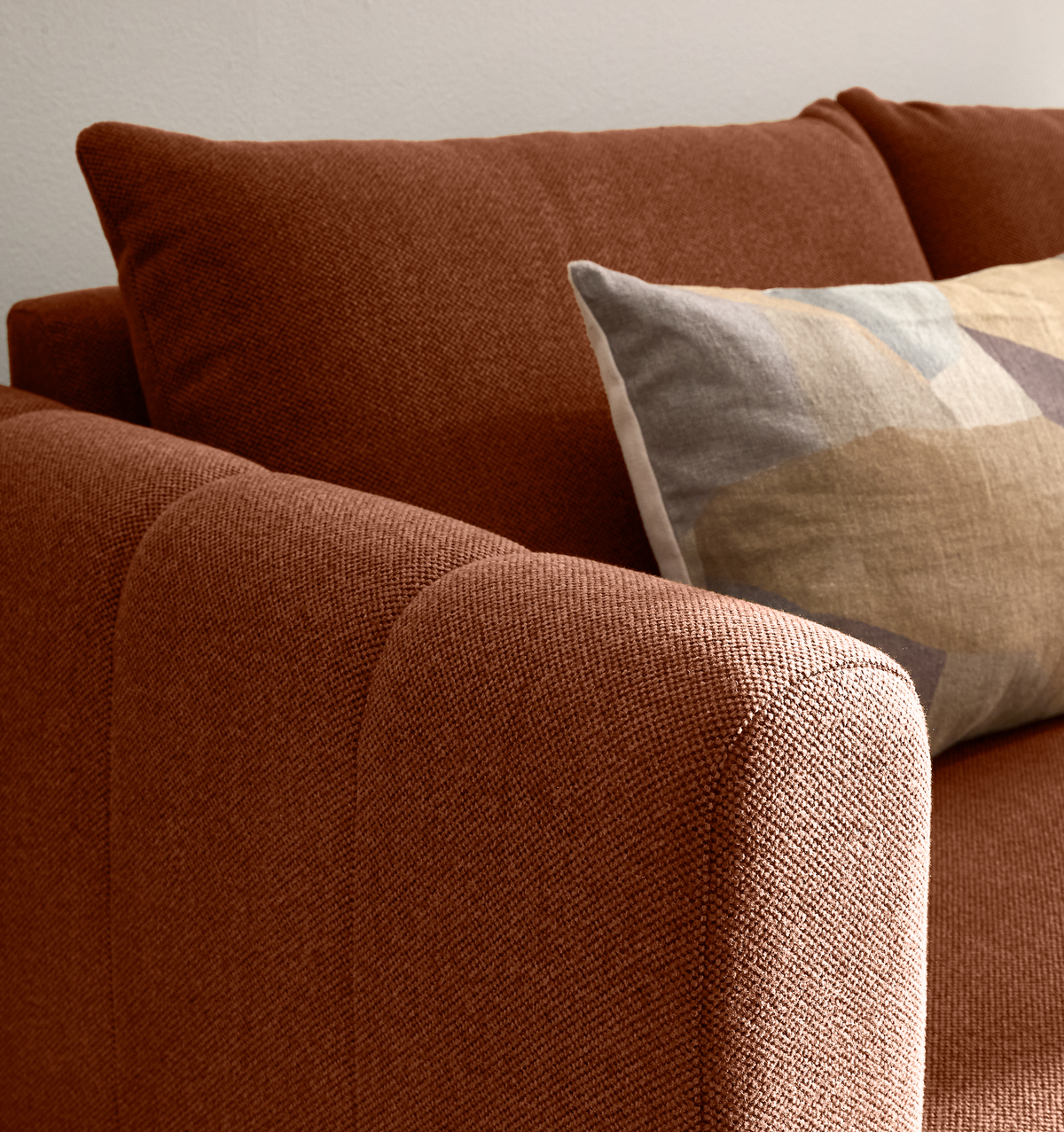 Nærbillede af en rustfarvet, tekstureret sofa med afrundet armlæn og en pude med geometrisk mønster i beige og grå.