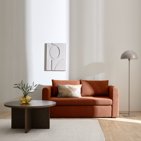 Rustrød sofa med pude, mørkt sofabord med gul glasvase og grøn kvist, vægkunst og kuppelformet gulvlampe i et lyst rum med solskygger.