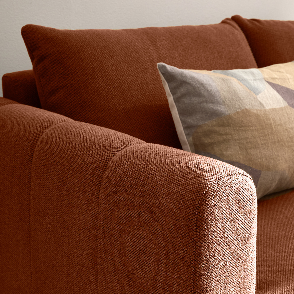 Nærbillede af rustfarvet stofsofa med afrundet armlæn og mønstret pude i beige og grå nuancer i dagslys.