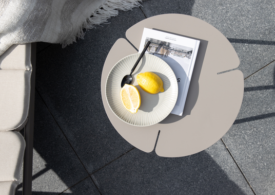 Rundt beige sidebord udendørs med tallerken, ske og citroner oven på en bog, solbelyst på fliser ved siden af en polstret bænk med tæppe.