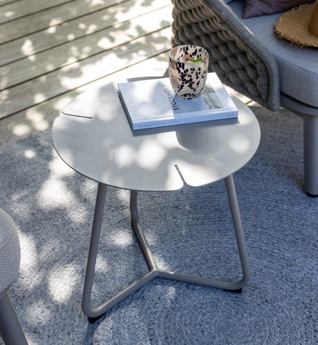 Rundt sidebord på terrasse med mønstret kop og bog ”STOCKHOLM” ovenpå, ved siden af grå flettet sofa på gråt tæppe i blødt dagslys.
