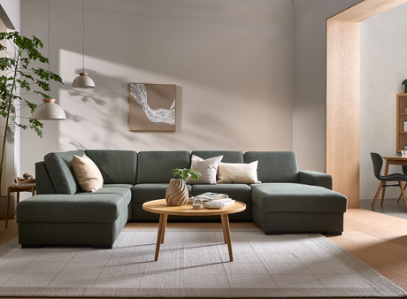 Stue med mørkegrøn hjørnesofa, lyse puder og ovalt sofabord i træ med vase og bog; to pendellamper, vægkunst og stort lyst tæppe i neutrale nuancer.